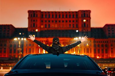 Man with arms outstretched on car sun roof in city at night