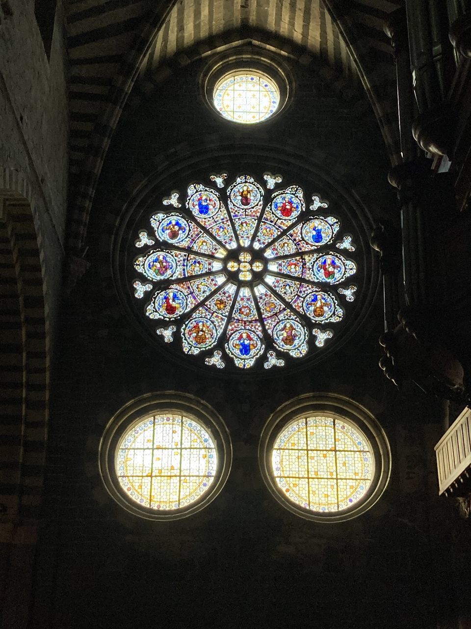 architecture, built structure, stained glass, indoors, glass, place of worship, window, low angle view, no people, light, ceiling, belief, religion, spirituality, building, circle, shape, geometric shape, lighting, catholicism, pattern, sunlight, ornate, travel destinations, arch, rose window