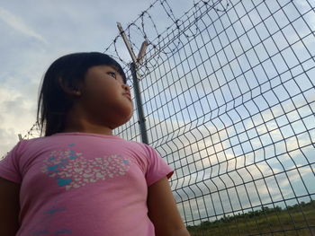 Girl looking away while standing on fence against sky