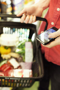 Midsection of man holding mobile phone while carrying grocery basket in supermarket