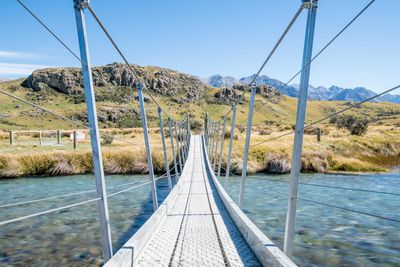 Empty footbridge over river against mountains and blue sky