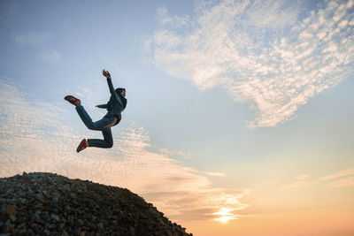 Low angle view of young woman jumping on rock against sky during sunset