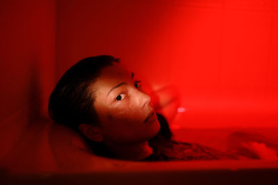 Bathroom portrait in red neon light of young sensual asian woman wearing clothes relaxing in bathtub