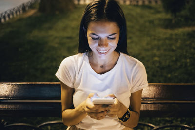 Happy woman using smart phone sitting on park bench at night