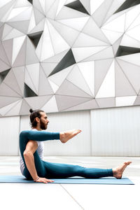 Side view of adult bearded man keeping leg on shoulder while doing stretching yoga exercise against wall with geometric ornament