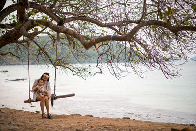 Portrait of woman sitting on swing while holding baby by sea