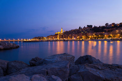 Illuminated buildings by sea against sky at dusk