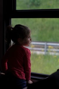 Rear view of girl looking through window while standing at home