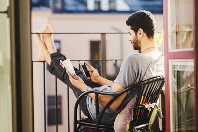 Side view of man using smart phone while holding guidebook at balcony