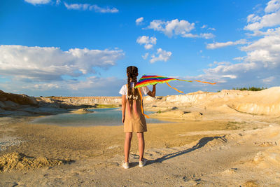 A girl with a kite stands on the edge of a mountain and looks into the distance, 