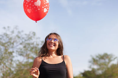 Smiling mid adult woman holding red helium balloon against sky