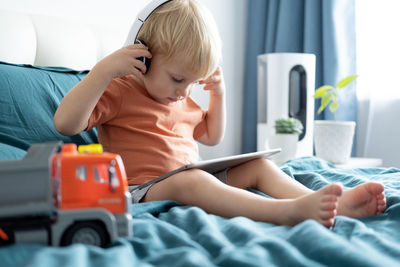 Smiling happy little kid boy using tablet in wireless headphones in bed at home