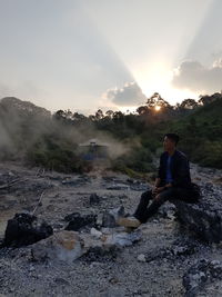 Man sitting on rock at shore against sky during sunset