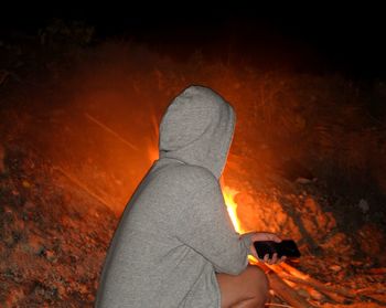 Side view of woman wearing hooded shirt crouching by campfire at night
