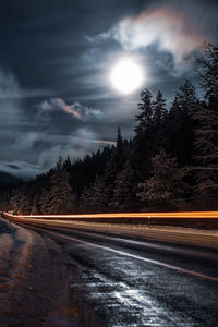 Light trails on road by trees against sky at night