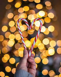 Hand holding two candy canes in heart shape with lights in background.