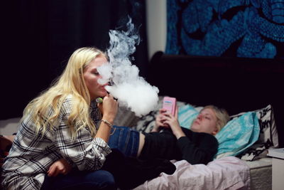 Woman exhaling smoke by sister on bed at home