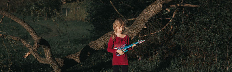 Man holding guitar standing by tree trunk in forest