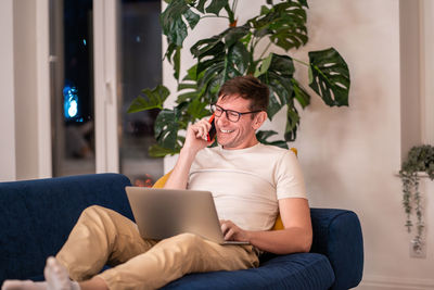 Laughing man in eyeglasses sitting on sofa with laptop on knees having fun cellphone conversation