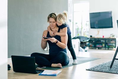 Daughter looking at smart phone while mother sitting on mat at home