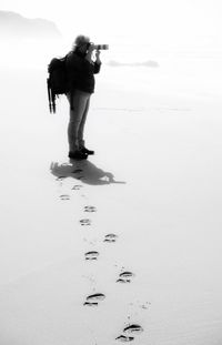 Side view of woman photographing while standing at beach against sky