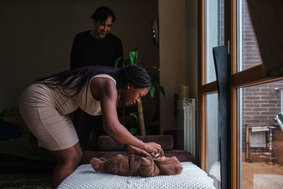 Side view of african american mother with braided hair feeding little infant in bear overall near ethnic attentive dad at home