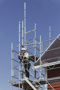 Full length female building contractor using smart phone standing at scaffolding