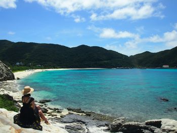 Rear view of woman sitting on rock at beach against cloudy sky