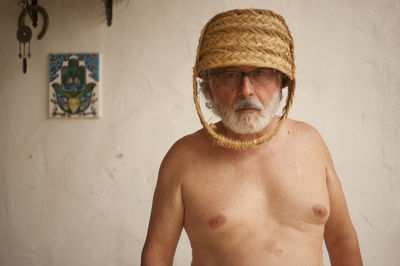 Shirtless man wearing basket on head while standing against wall