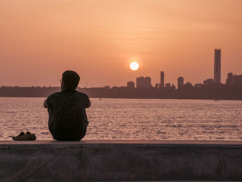 Rear view of silhouette man sitting by river against sunset sky