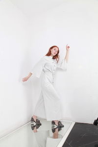 Full length fashion catalog style portrait of woman in jumpsuit standing on mirror over white wall