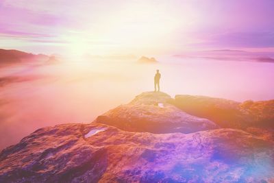 Alone man on rocky cliff overlooking misty plains below at border of valley. abstract light flare