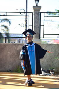 Portrait of girl wearing graduation gown and mortarboard while standing against wall