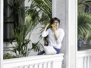 Woman speaks on smartphone.  female in eyeglasses sits on balcony with palm trees in flower pots.