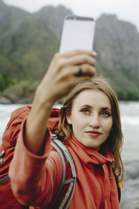 Female hiker taking selfie with mobile phone while standing at riverbank against mountain