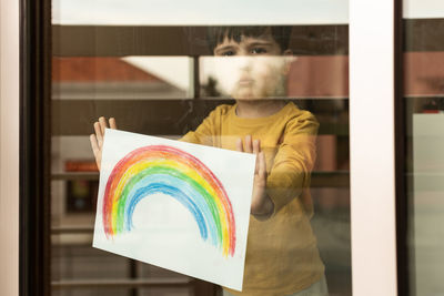 Portrait of boy holding multi colored rainbow painting seen through window