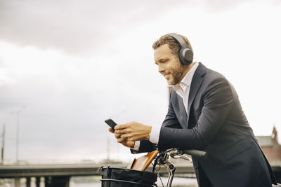 Businessman using mobile phone while leaning on bicycle handle against sky