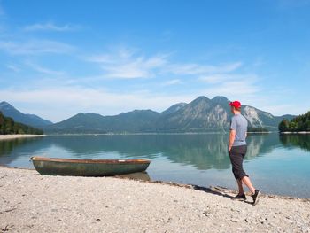 Tall man walks at bench on lake shore and looks at the sports boat on the level.