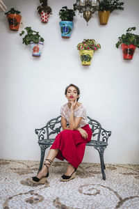 Graceful ethnic female in red skirt and lips sitting on stool in patio of house in summer while touching neck and looking at camera