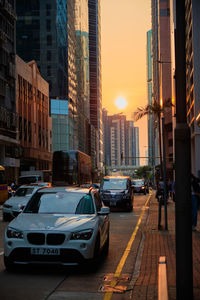 Traffic on city street by buildings against sky during sunset