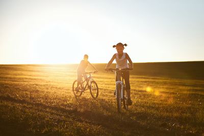 Siblings riding bicycle on field against sky during sunset