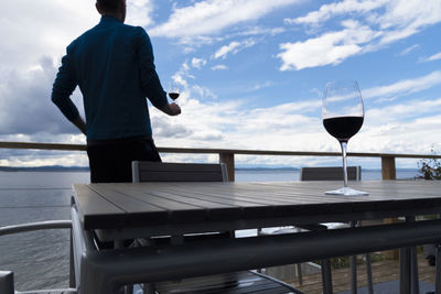 Rear view of man holding wineglass by sea against sky