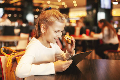 A cute schoolgirl sits in a cafe, plays on a tablet and eats a hamburger. happy child eating