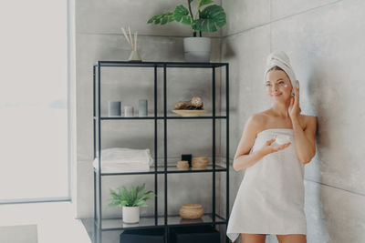 Young woman applying cream on face while standing by wall