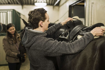 Young man preparing horse in stable with woman in background