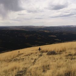 Rear view of person standing amidst dry grass on mountain against cloudy sky