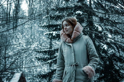 Woman standing while looking away against snow covered trees in forest