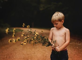 Shirtless boy holding flowers standing on dirt road