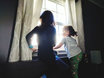 Siblings looking through window while sitting at home on sunny day