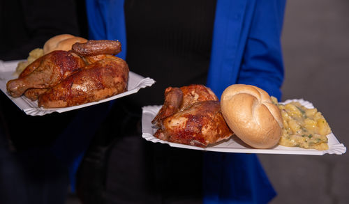 Plates with grilled chicken, bread rolls, and potato salad, held by people, close-up view